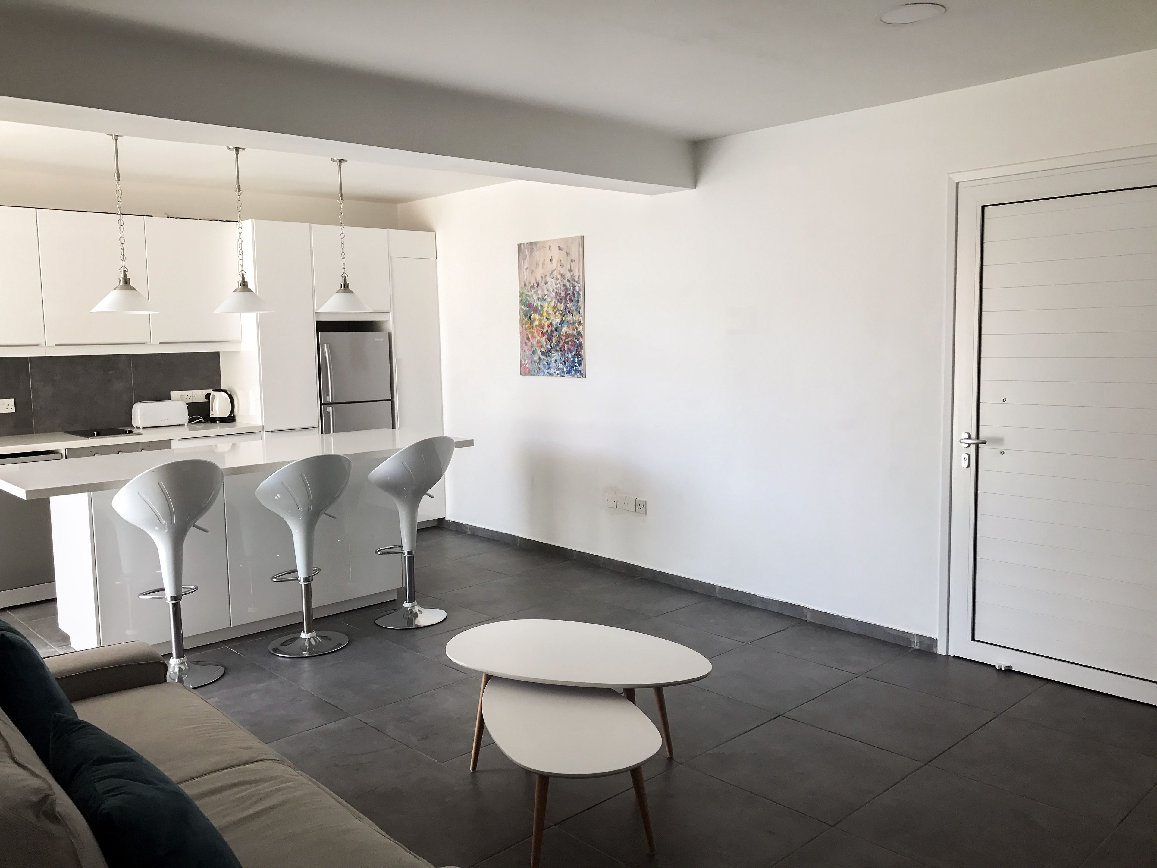 A modern kitchen with white cabinets, a stainless steel refrigerator, and pendant lighting. There is a dining area with a white table and two bar stools.