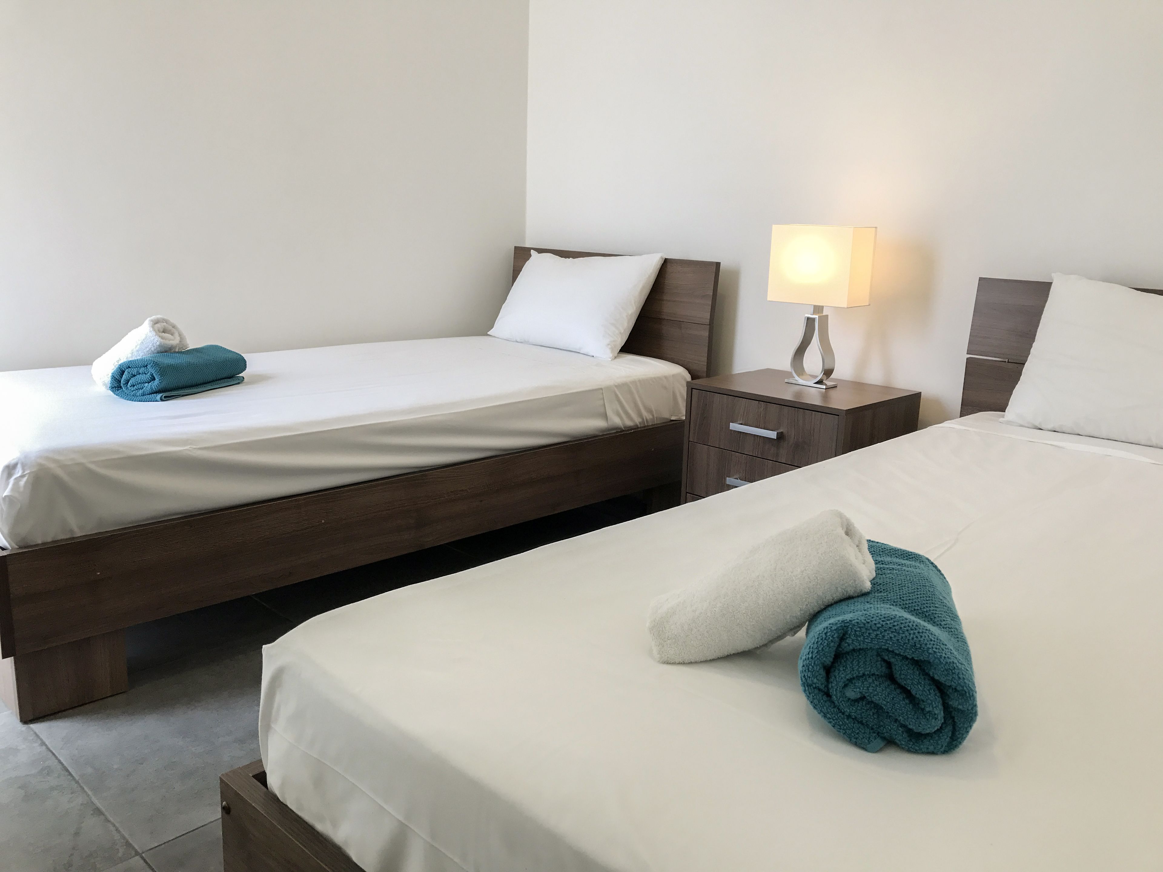 Une chambre d’hôtel avec deux lits, chacun doté de draps blancs et d’une tête de lit en bois. Une table de chevet avec une lampe se trouve entre les deux lits.