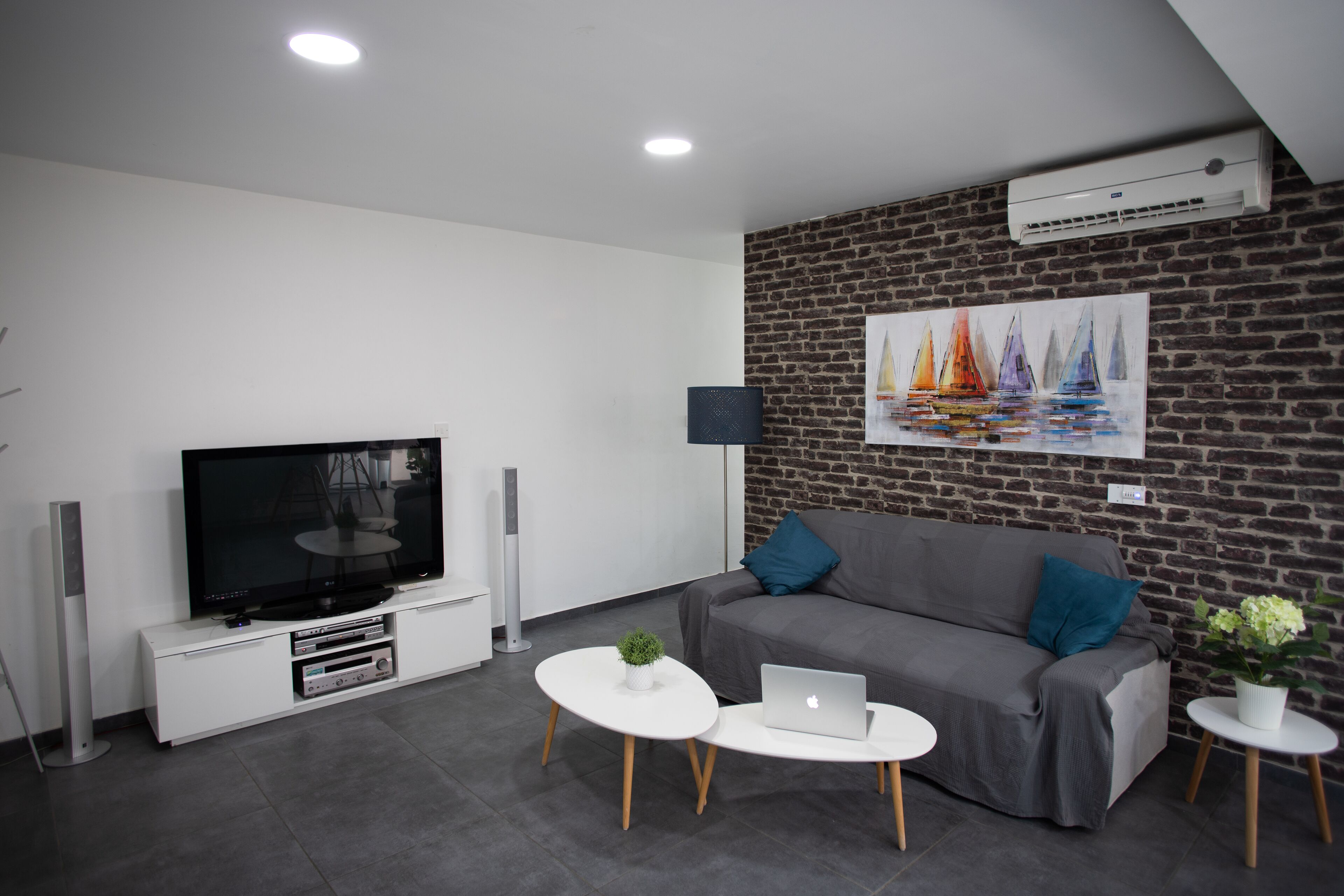 Un salon moderne doté d’un téléviseur à écran plat, d’un canapé gris, de deux tables basses blanches et d’un mur au motif de briques orné d’un tableau.