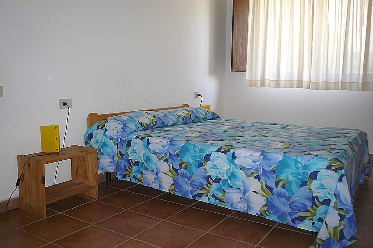 Une chambre à coucher avec un lit, une table de chevet en bois, une prise électrique au mur et une fenêtre avec des rideaux.