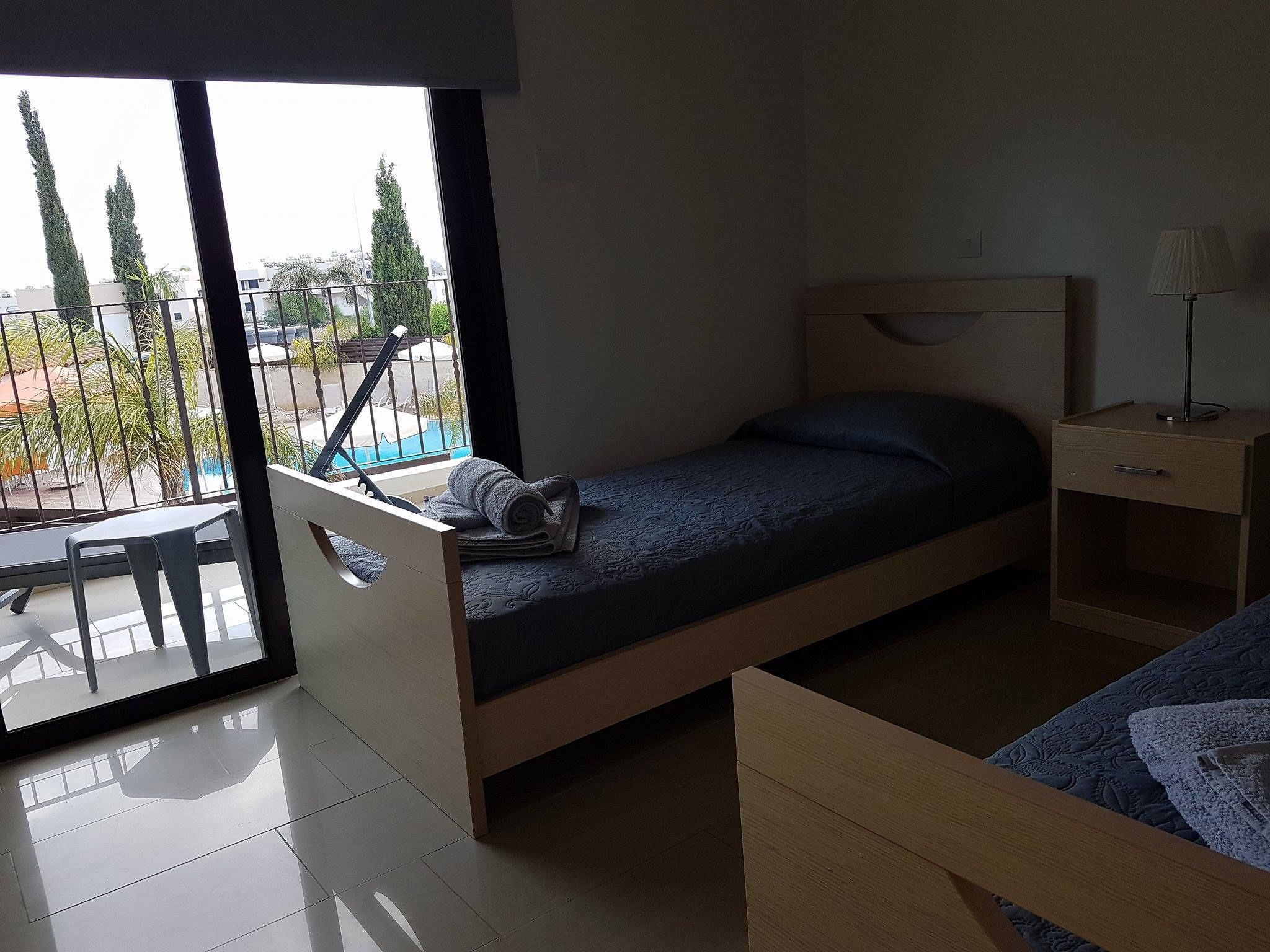 Un dormitorio moderno y pequeño con una cama individual, una mesita de noche con lámpara y vistas a un balcón con mobiliario exterior.