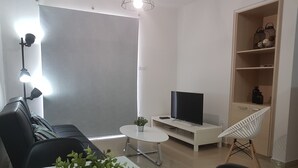 A modern living room with a sofa, a TV on a white stand, a small table with a plant, a chair, and a shelf with decorative items. - Marand Apartment BF11 (Paralimni)