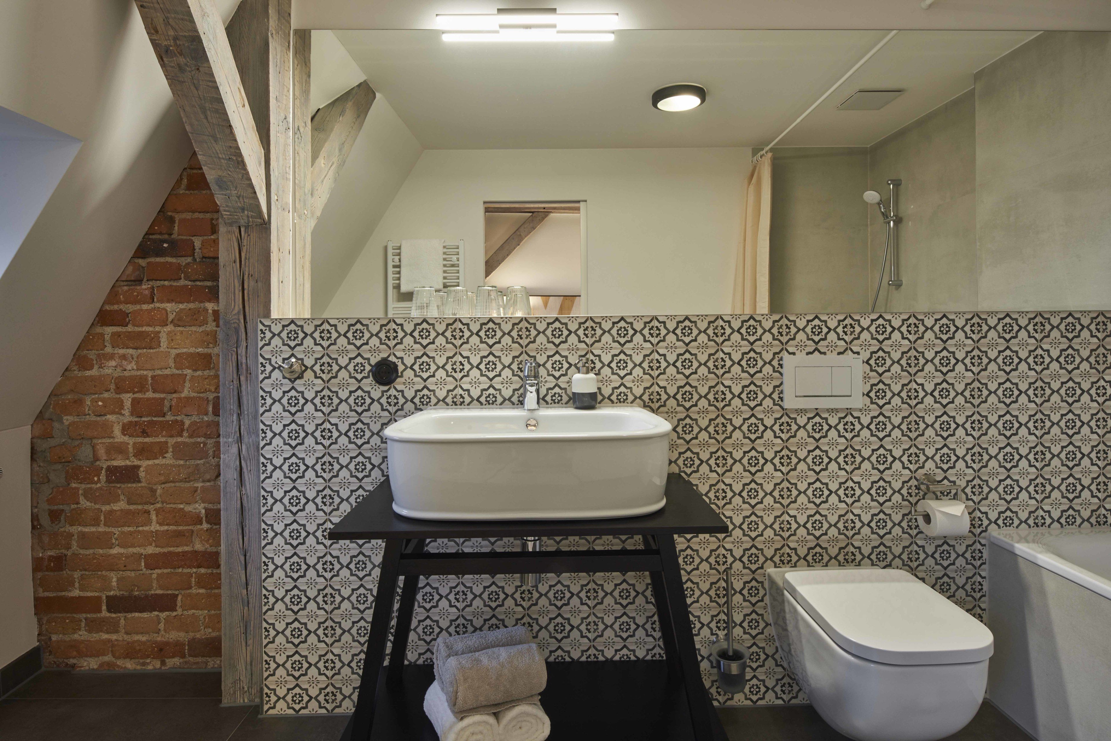Een badkamer met een wit wastafel, toilet en douche. Een wand met een patroon en een bakstenen muur aan de zijkant.