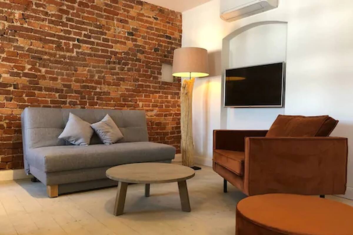 Un salon avec un mur en briques, un canapé gris, un fauteuil marron, une table basse ronde et un lampadaire.