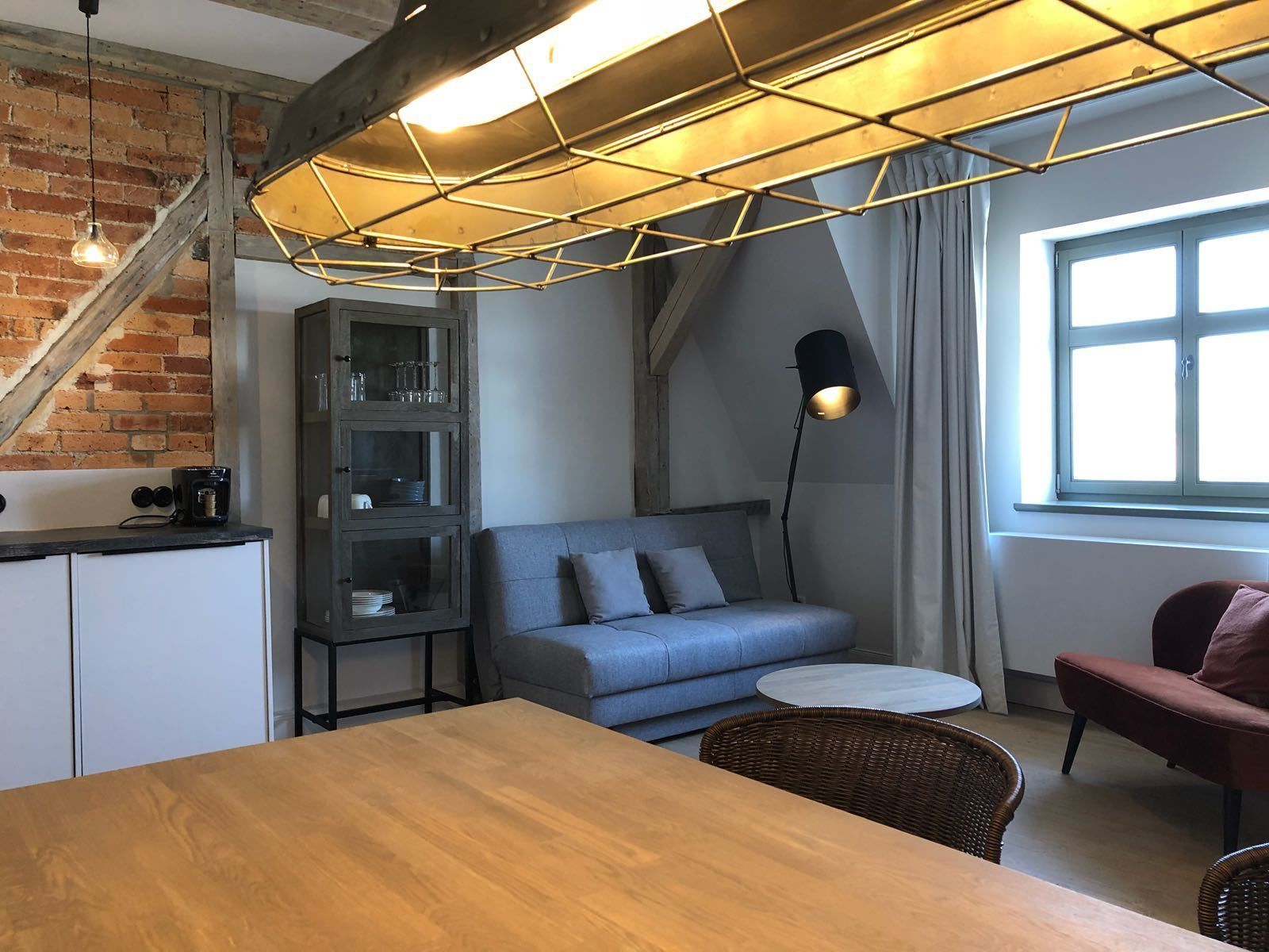 Una sala acogedora con una mesa de madera, un sofá azul, un sillón rojo, una estantería y una escalera con paredes de ladrillo visto.