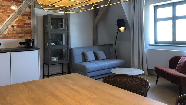 Ein gemütlicher Wohnbereich mit einem Holztisch, einem blauen Sofa, einem roten Sessel, einem Bücherregal und einer Treppe mit freiliegenden Backsteinwänden.