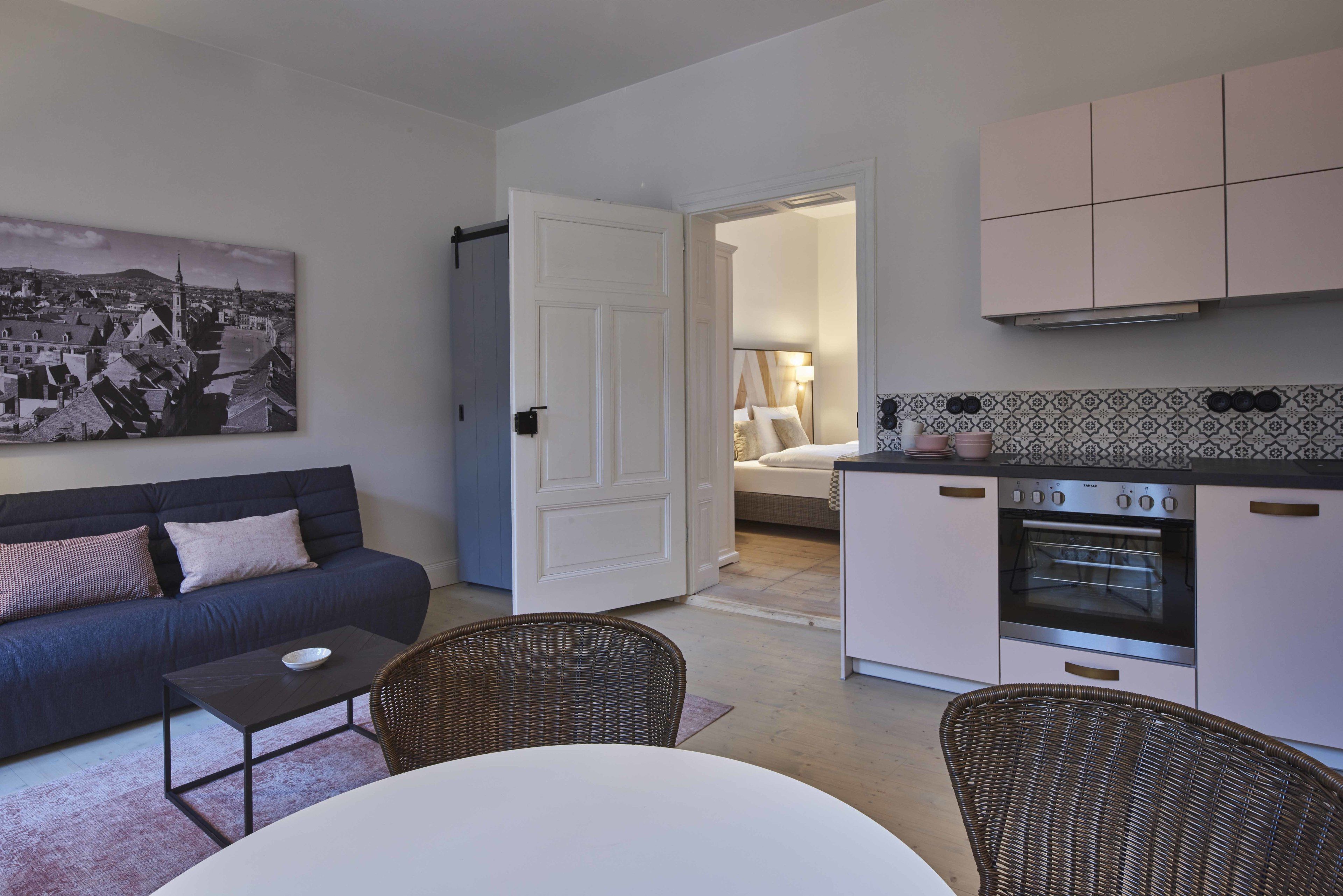 A compact living space with a sofa, dining table, wicker chairs, a kitchen with an oven, and a bedroom with a bed and nightstand.