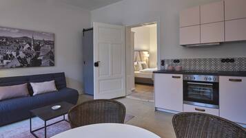 Um espaço compacto com sofá, mesa de jantar, cadeiras de vime, cozinha equipada com forno e quarto com cama e mesa de cabeceira.