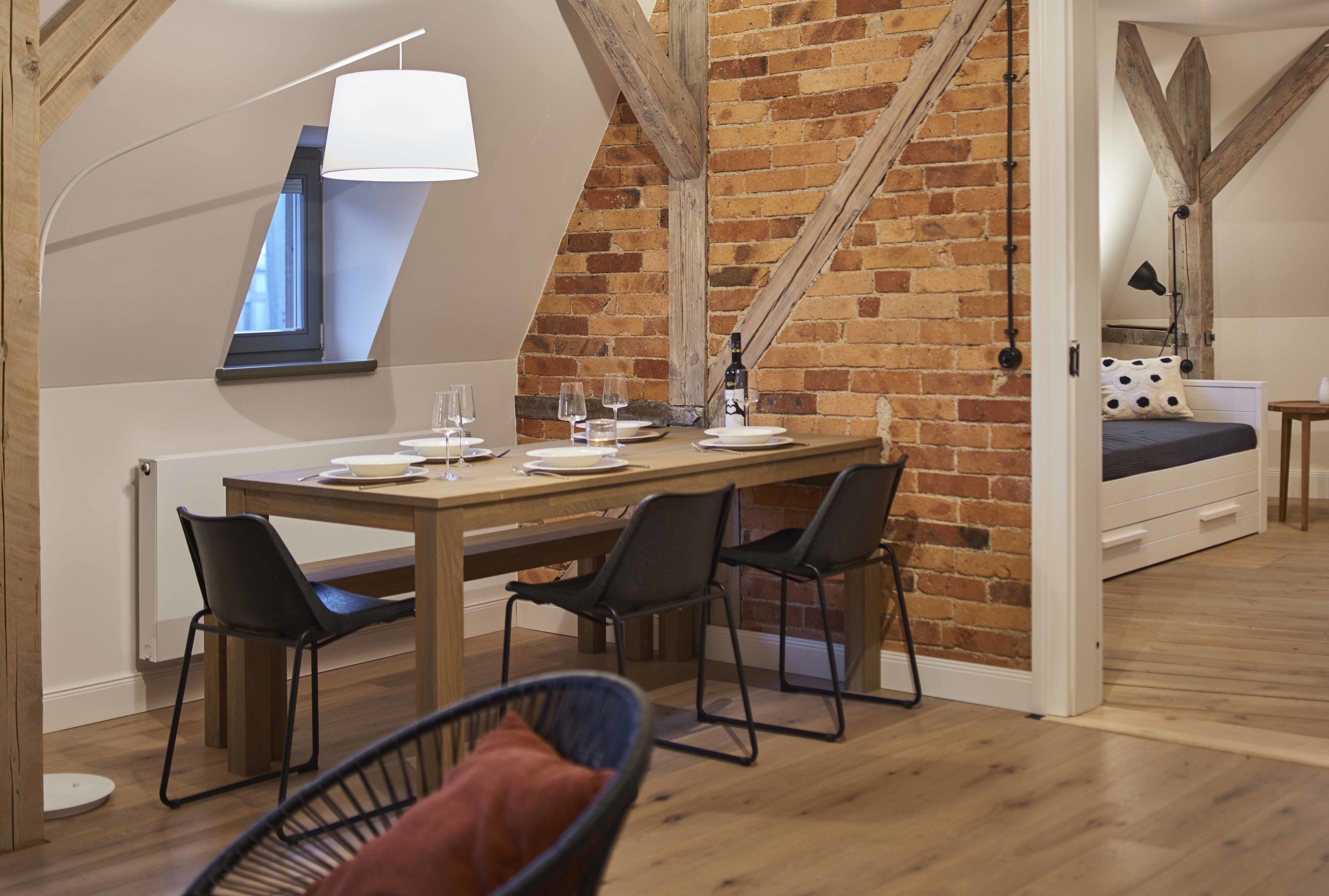 A dining area with a wooden table, four chairs, and a bench. A brick wall and wooden beams are visible in the background.