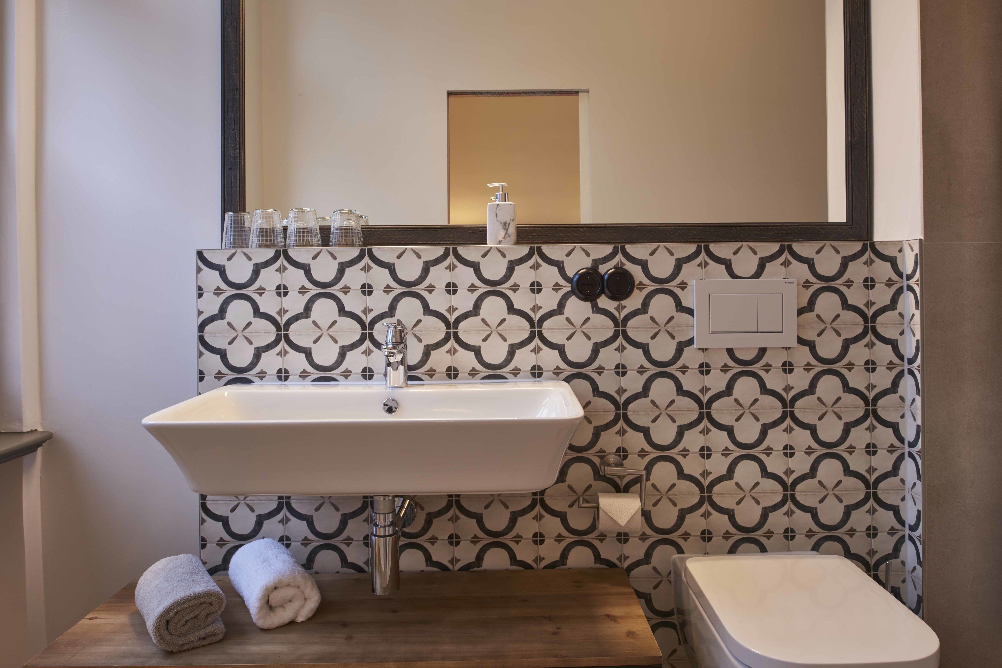 Un baño con un lavamanos blanco, un espejo encima y baldosas con diseño en la pared.