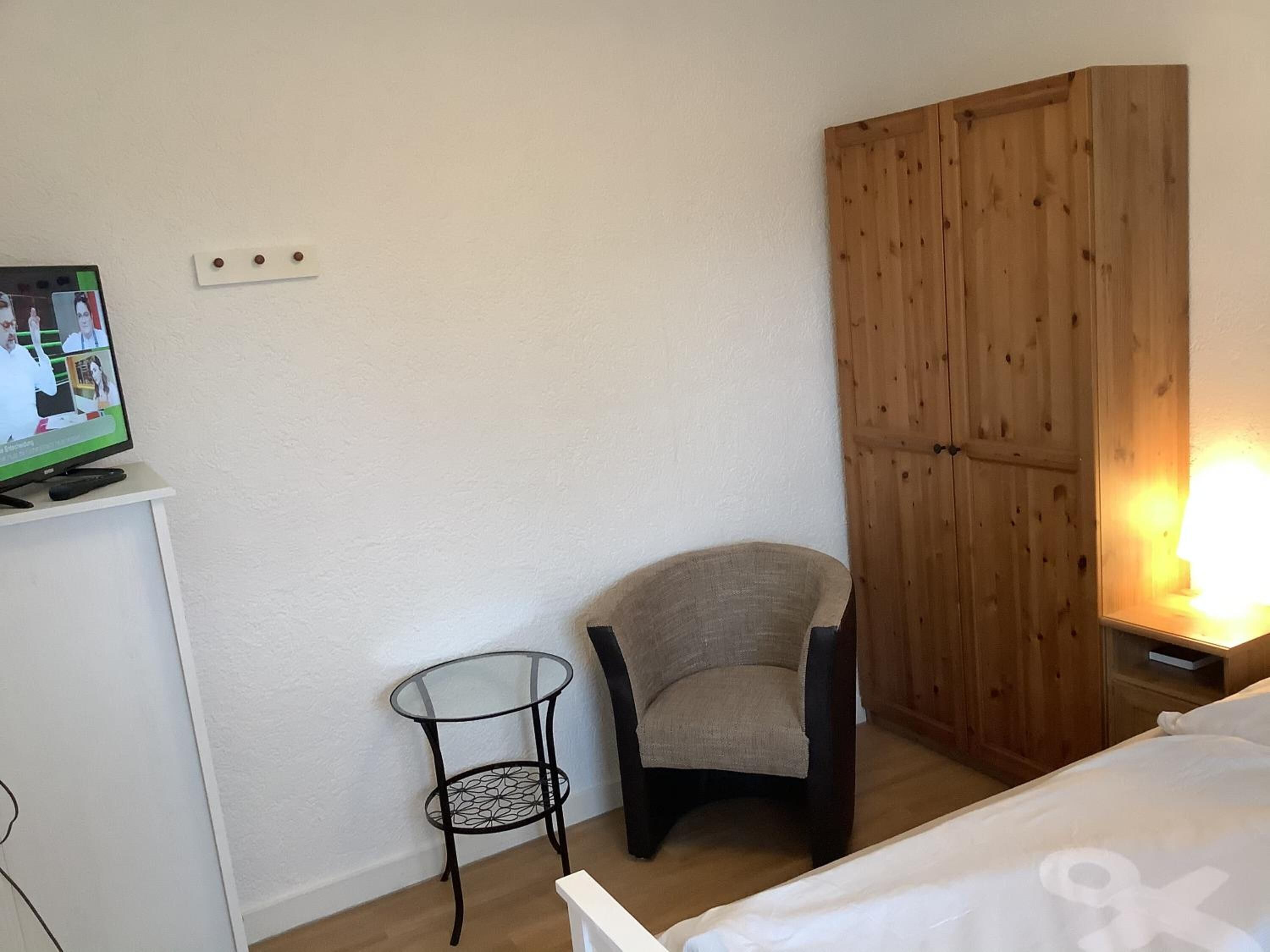 Quarto com uma cama, uma cadeira, uma mesa pequena, um guarda-roupa de madeira e uma televisão sobre um suporte.
