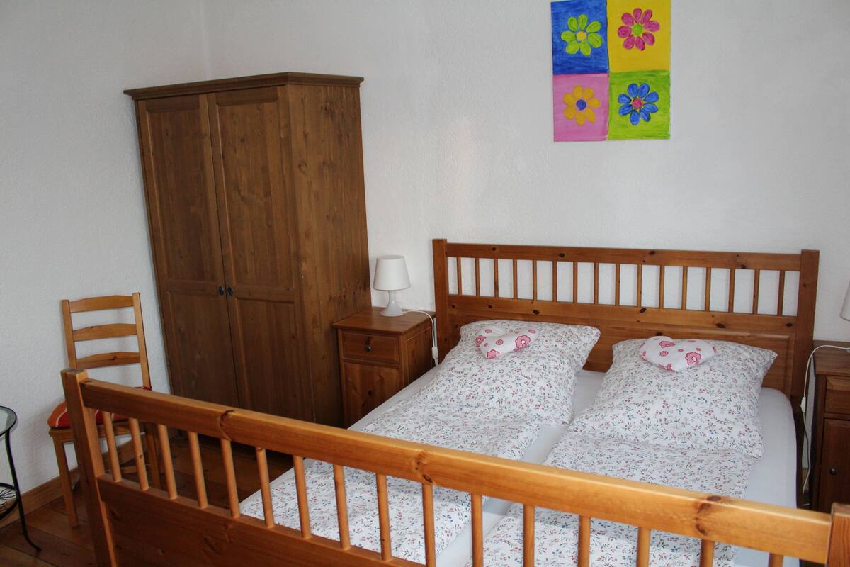 Een slaapkamer met een houten bed, een houten kledingkast, een nachtkastje met een lamp en een wand met kleurrijke bloemenkunst.