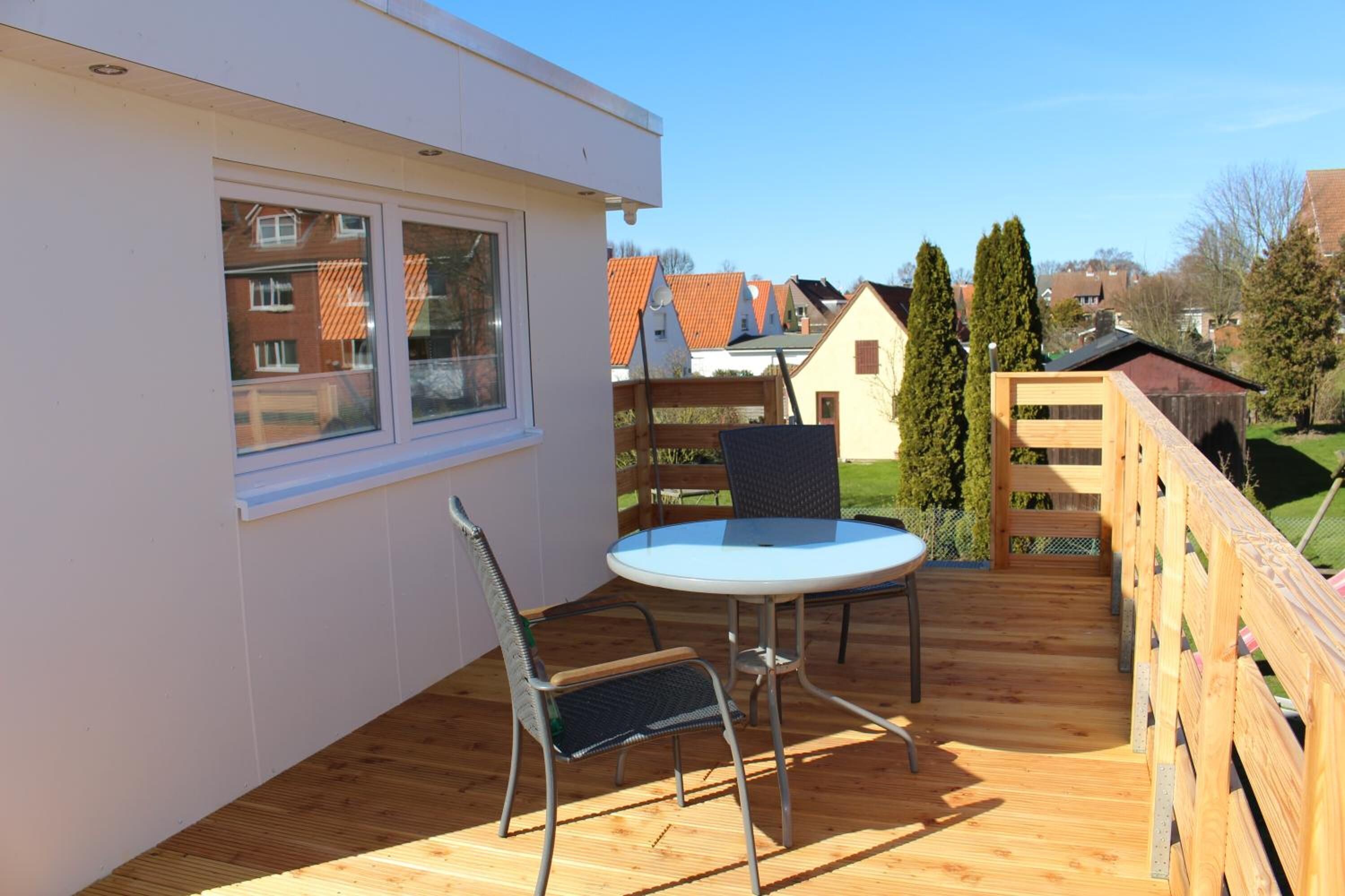 A wooden deck with a round table and two chairs, a window, and a view of neighboring houses.