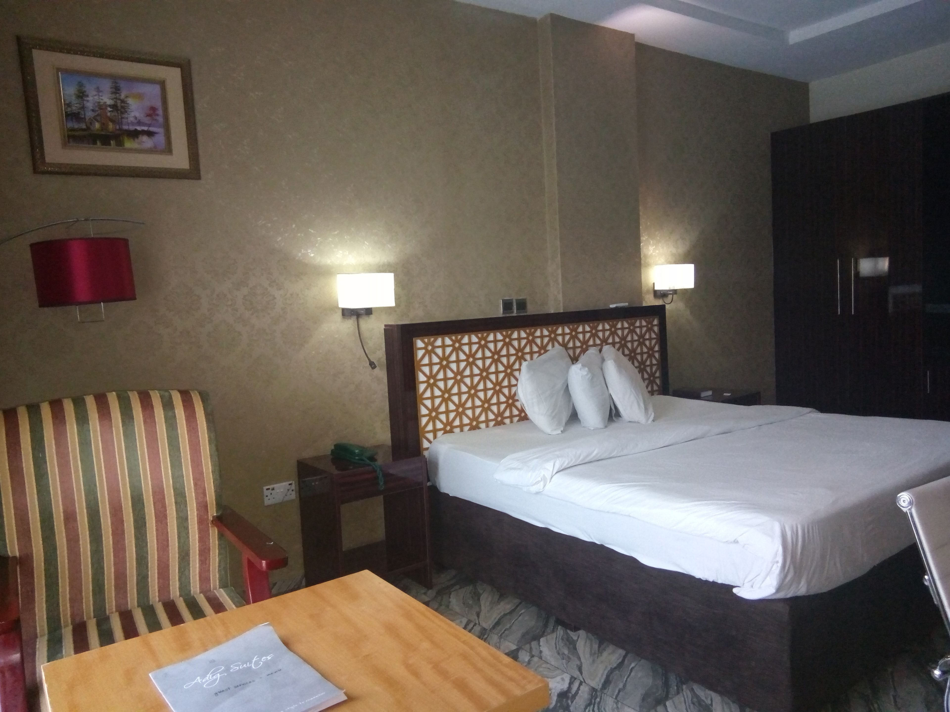 A hotel room with a bed, a striped chair, a small table with a piece of paper, and two bedside lamps.