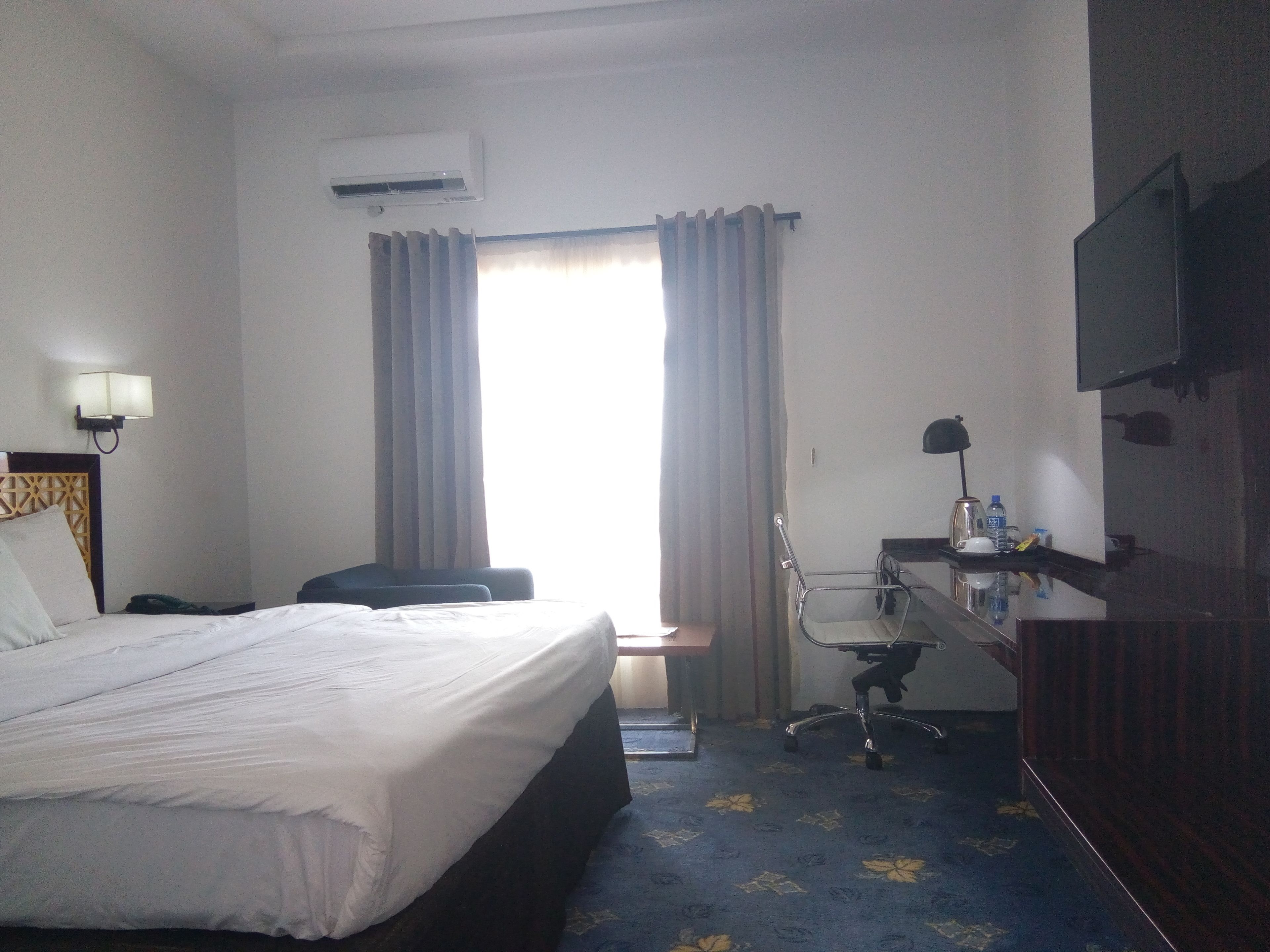 A hotel room with a bed, a desk, a chair, a TV, and an air conditioning unit.