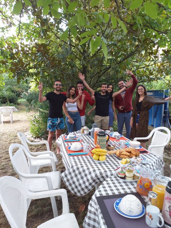 A group of people standing around a checkered table with food and drinks, outdoors with trees in the background. - Chambre du Jardin Fleuri (Saint-Aubin-Les-Elbeuf)