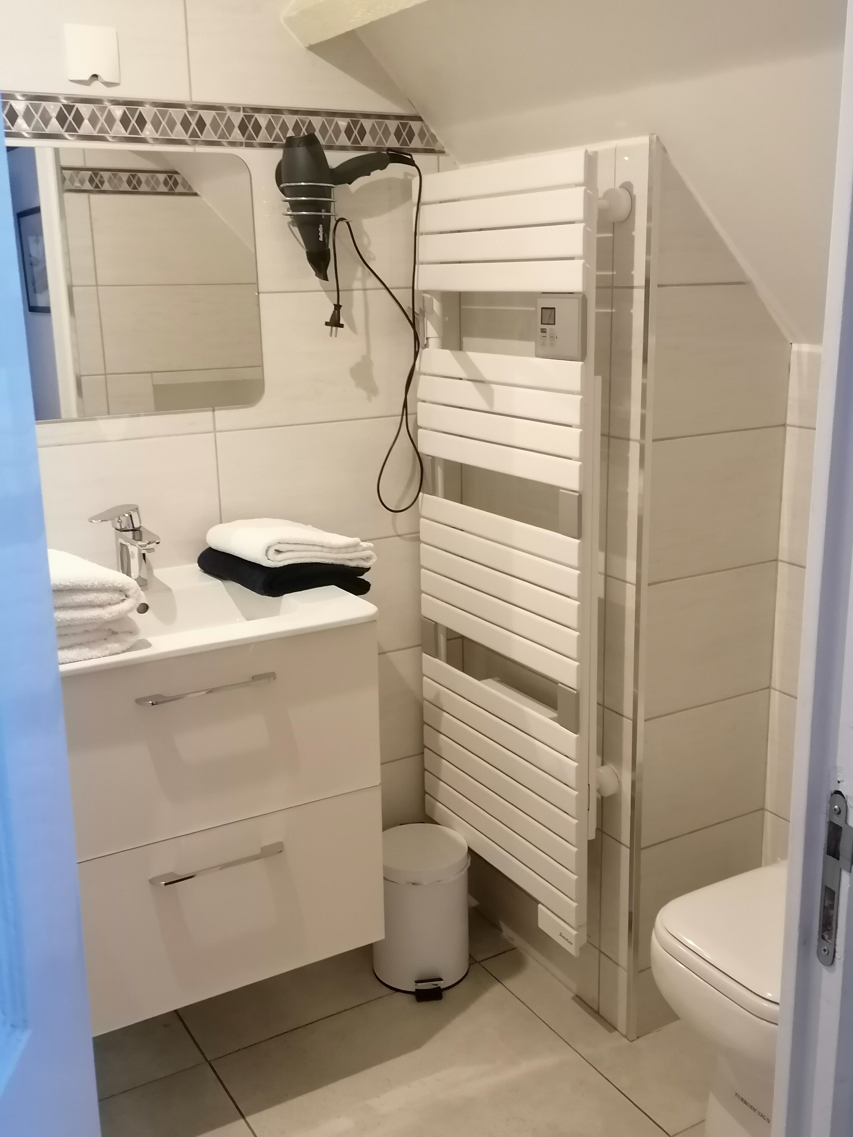 Une salle de bain avec un lavabo blanc, un miroir et un porte-serviettes. On y trouve également des toilettes blanches et une douche avec un porte-serviettes blanc.