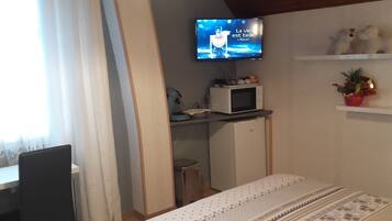 Habitación de hotel pequeña con una televisión mostrando un programa francés, microondas, refrigerador pequeño, mesa de comedor y una planta en maceta.