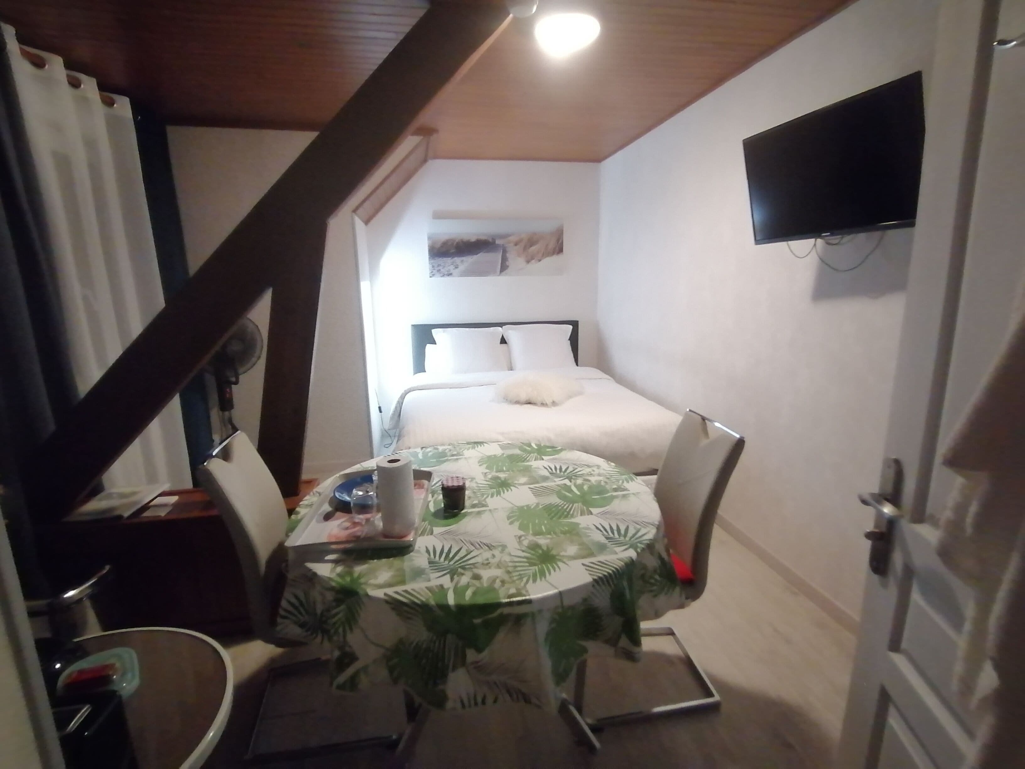 Une petite pièce comprenant un lit, une table recouverte d’une nappe, une chaise, un téléviseur fixé au mur et un ventilateur de plafond.