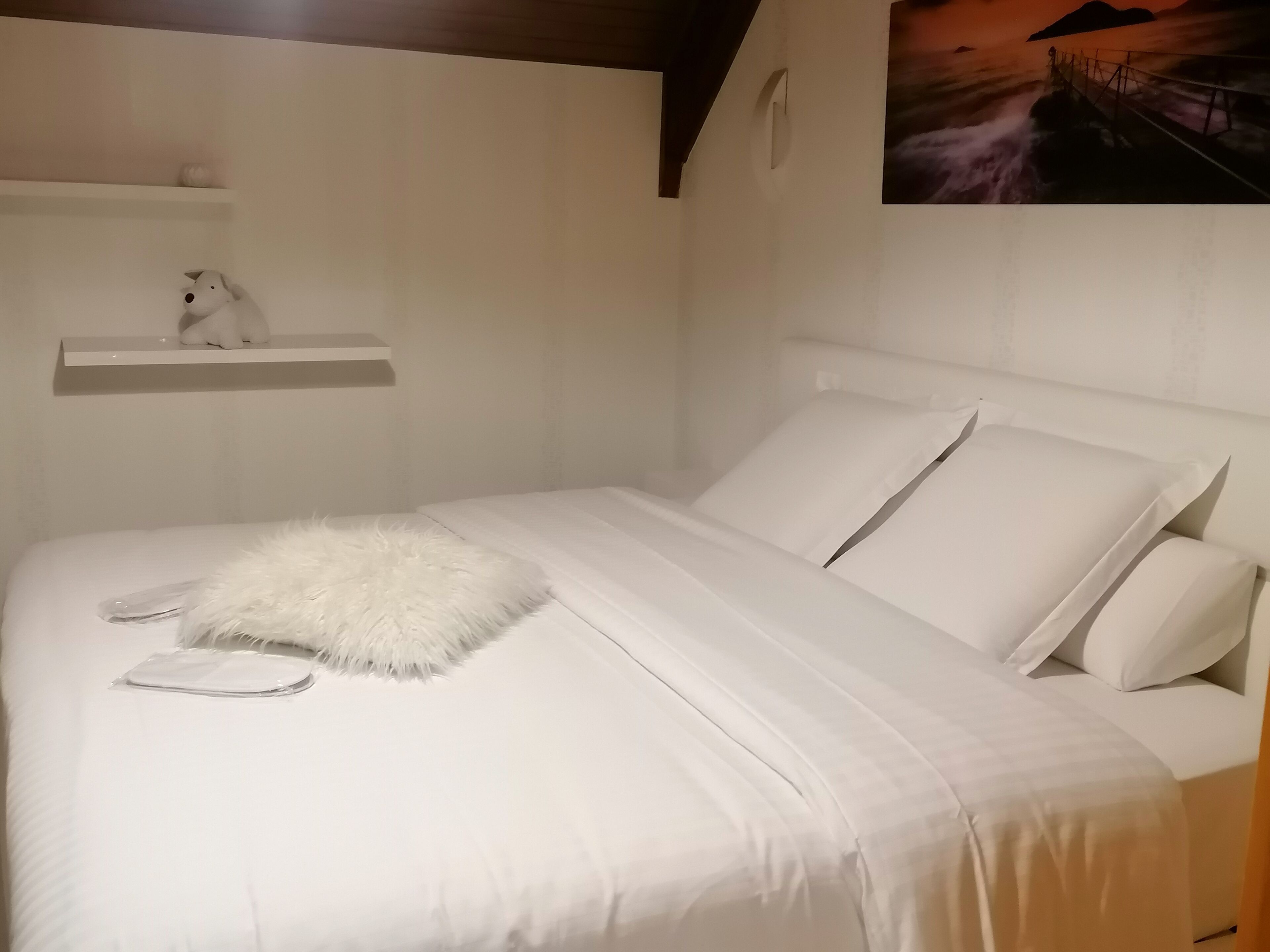 Un letto rifatto con lenzuola bianche e un soffice cuscino bianco, uno scaffale bianco con un piccolo oggetto decorativo, e un quadro di un ponte sull'acqua appeso al muro.