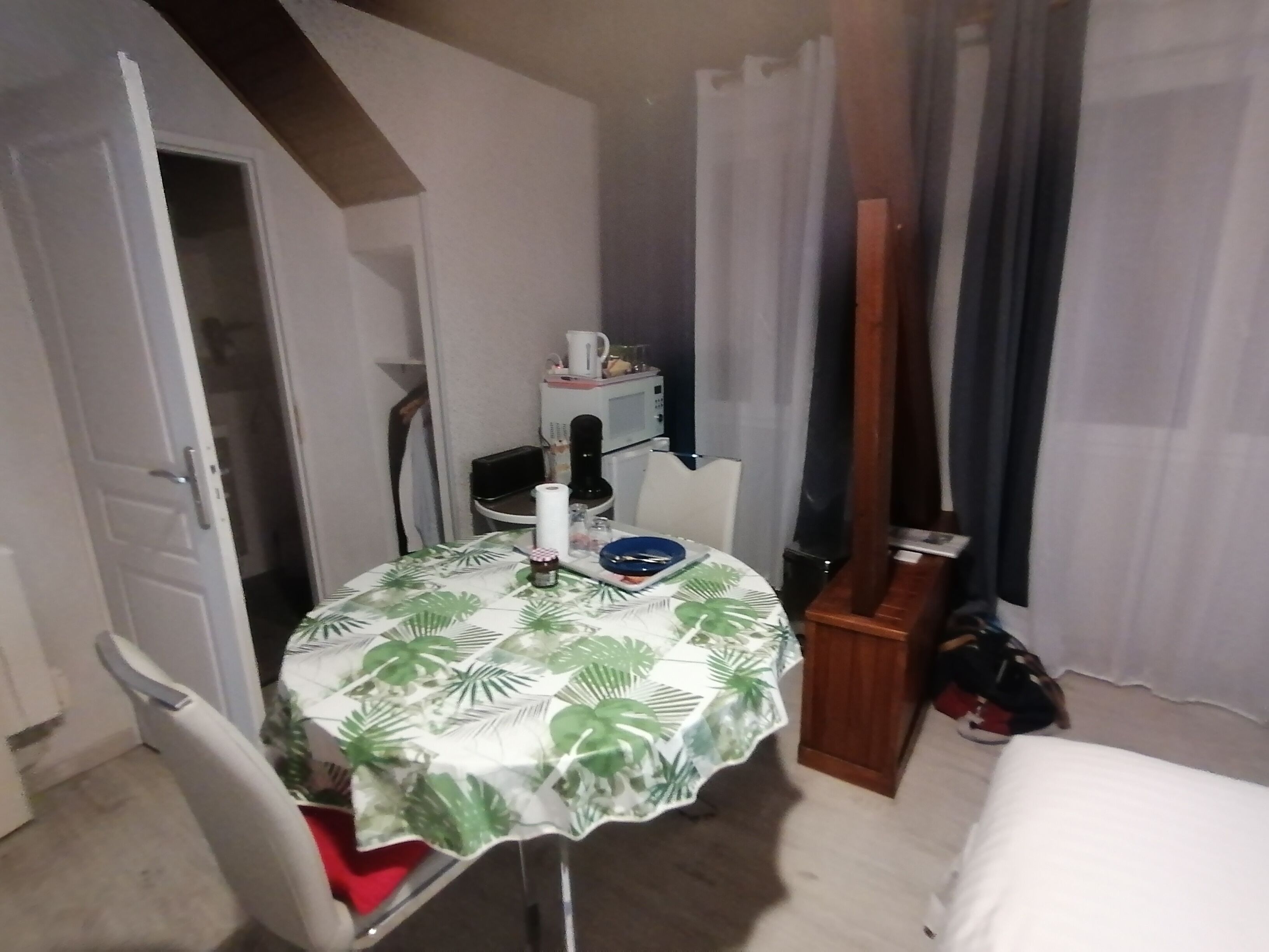 Pequeña habitación con una mesa de comedor cubierta por un mantel de hojas tropicales, una silla, un mueble pequeño y una cama.
