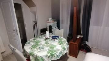 Pequeña habitación con una mesa de comedor cubierta por un mantel de hojas tropicales, una silla, un mueble pequeño y una cama.