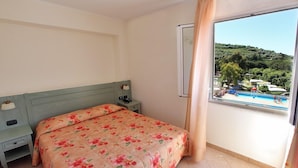 A bedroom with a bed, a nightstand, a window with a view of a pool and greenery. - Il Villaggio di Giuele (Finale Ligure)