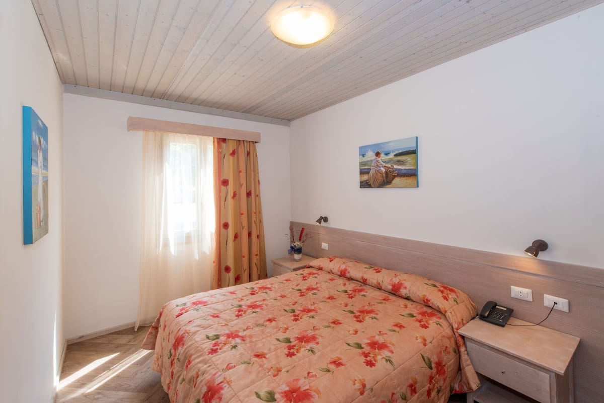 A bedroom with a bed, a nightstand, a lamp, a painting, and a window with curtains.