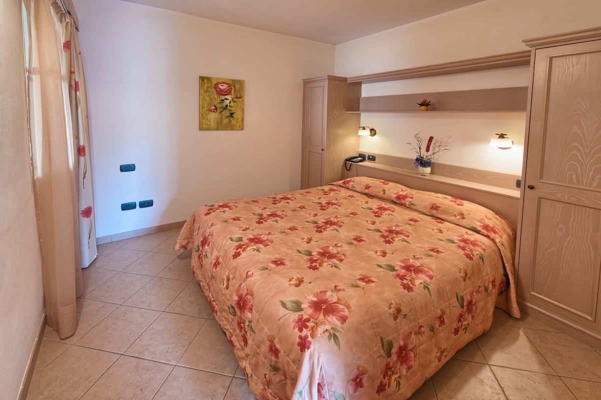 A bedroom with a floral bedspread, a nightstand with a lamp, a small shelf with decorative items, and a door with a wooden panel.