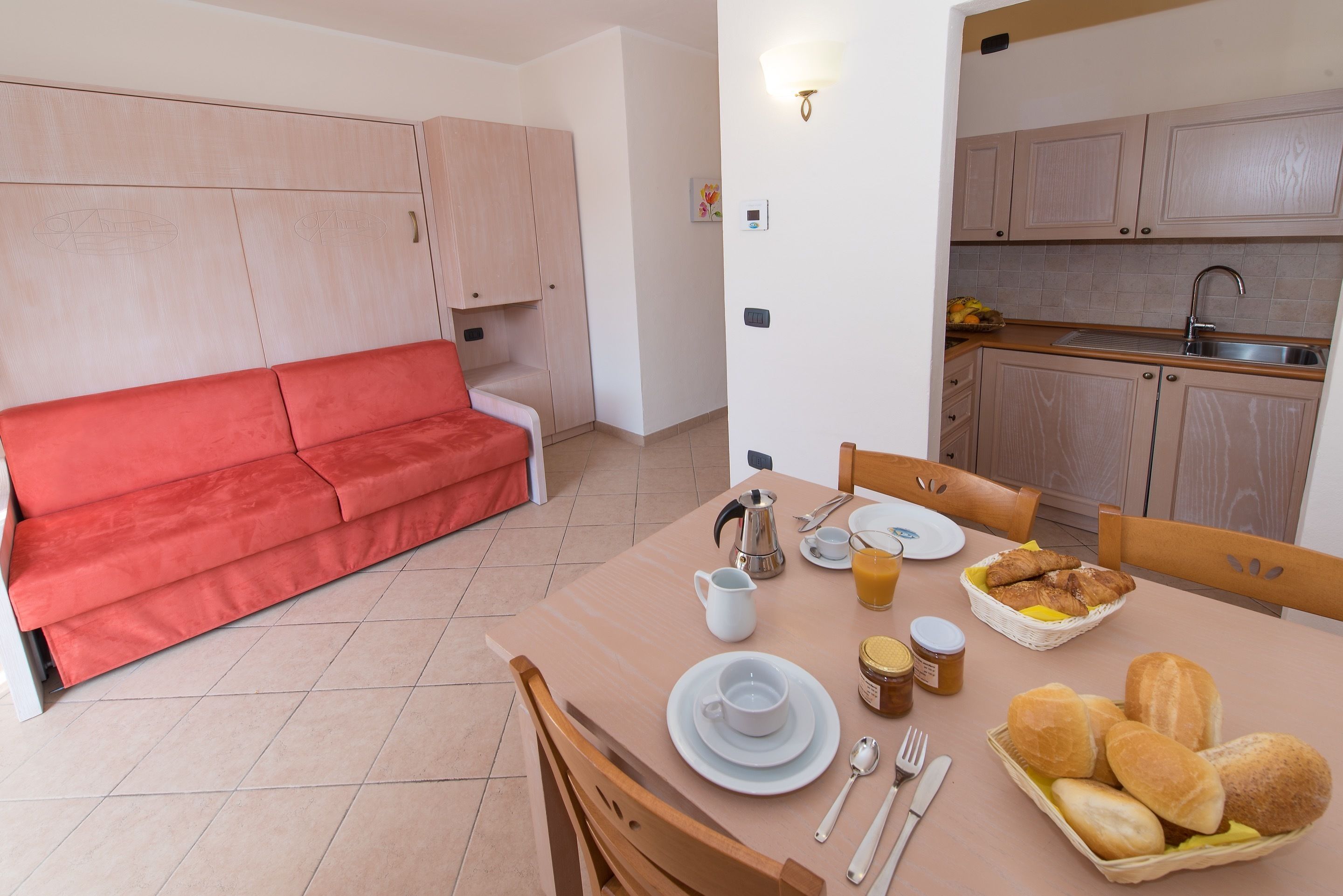 Un espacio habitable compacto con un sofá rojo, una mesa de comedor con provisiones para el desayuno y una kitchenette.