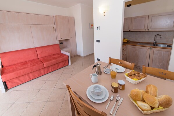 A compact living space with a red sofa, a dining table set with breakfast items, and a kitchenette. - Il Villaggio di Giuele (Finale Ligure)