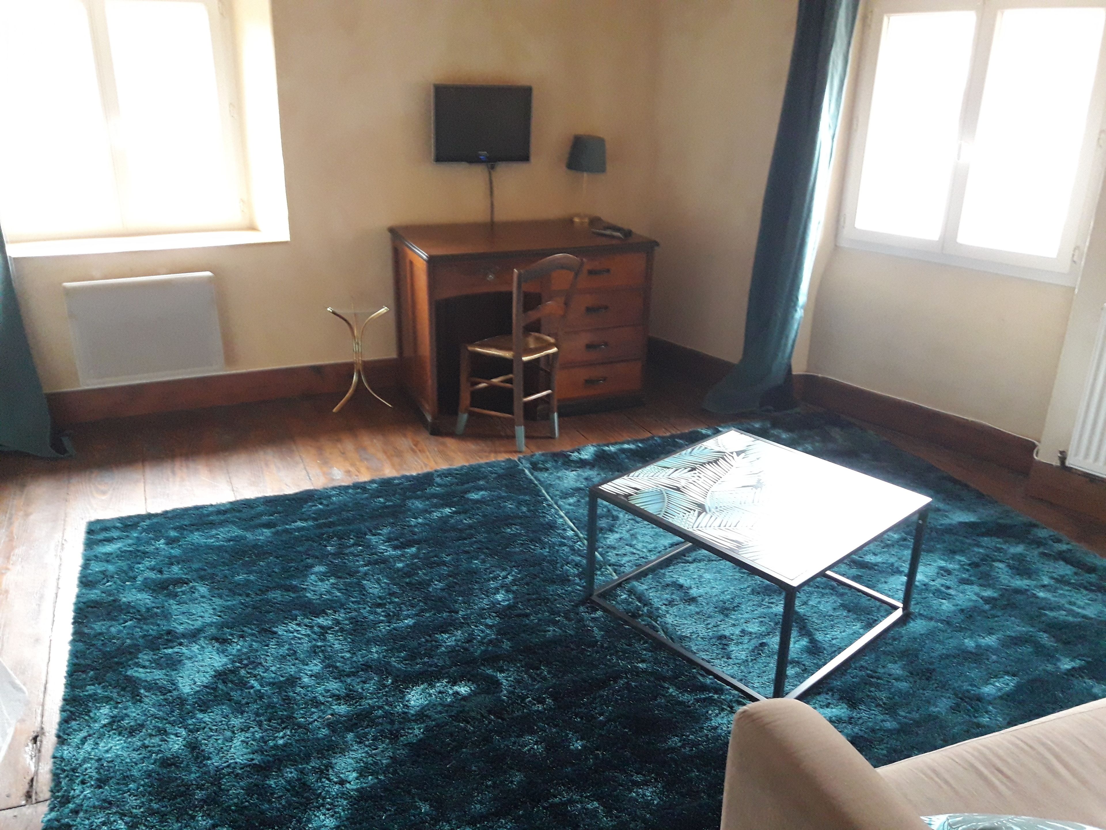 Una habitación con un escritorio de madera, una silla, una mesita pequeña, una alfombra azul y un televisor en la pared.