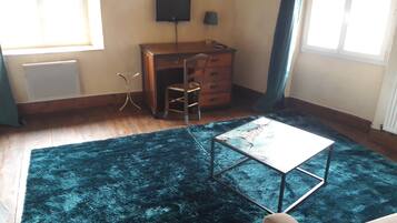 Una habitación con un escritorio de madera, una silla, una mesita pequeña, una alfombra azul y un televisor en la pared.