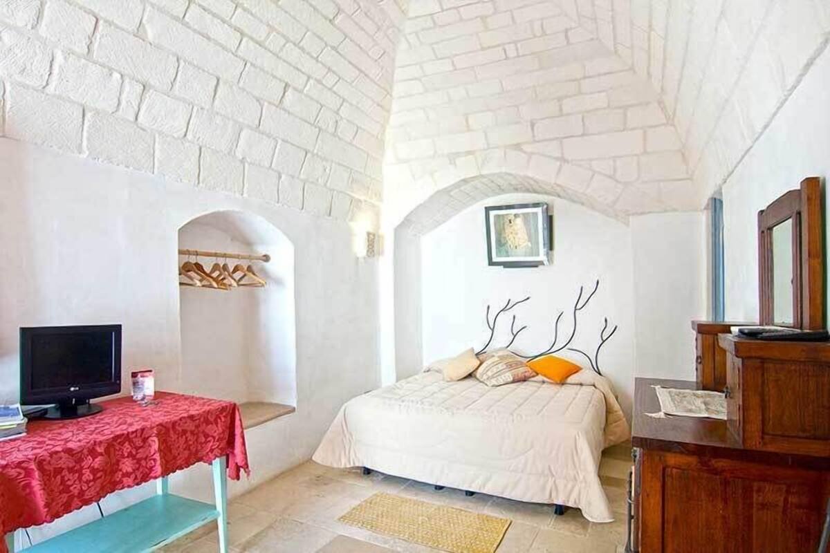 Un dormitorio con techo de ladrillo, una cama con cabecera blanca, un aparador de madera, un televisor sobre una mesita y un ventanal pequeño.