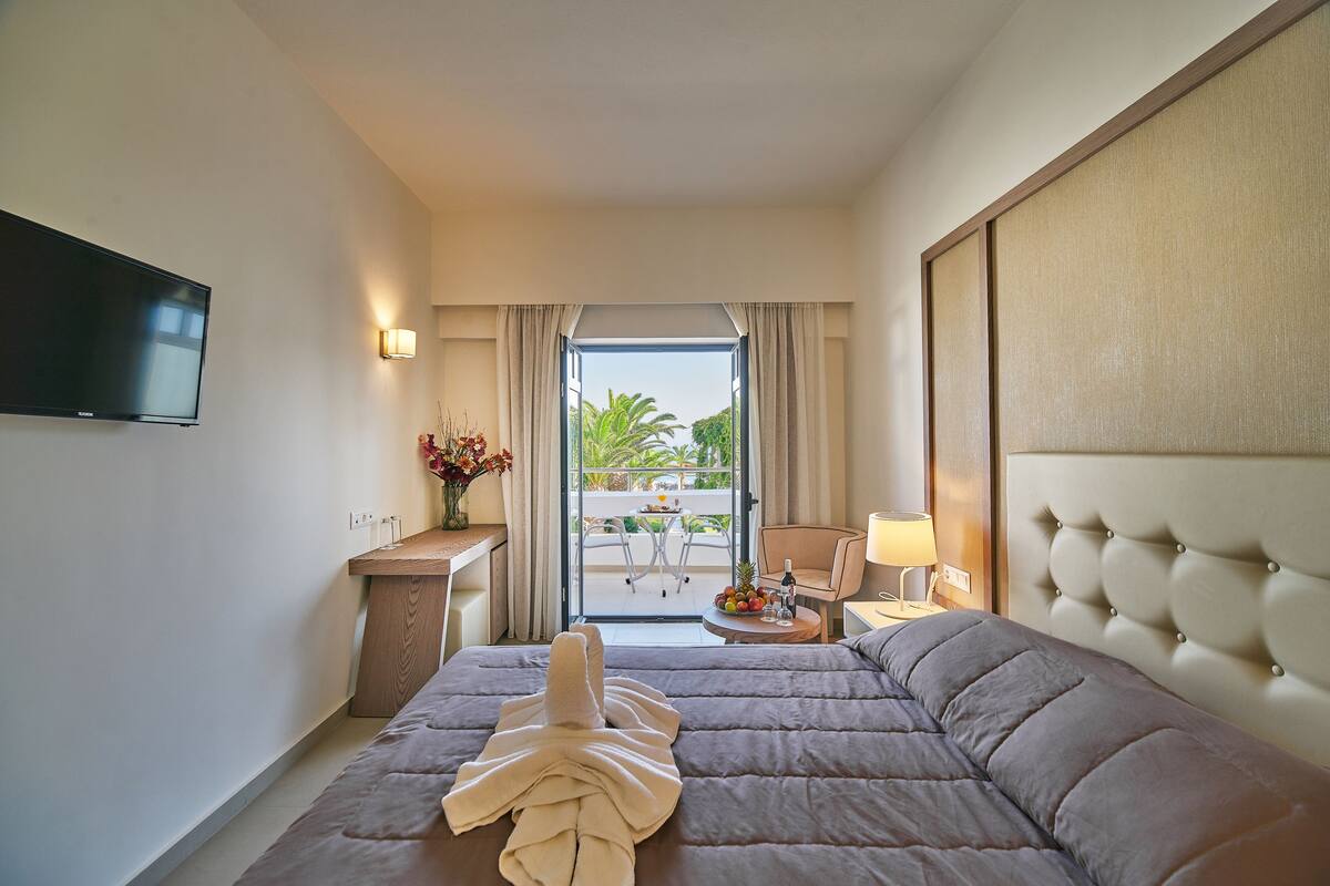 A hotel room with a bed, a TV, a desk, a chair, a small table with fruit and a bottle, and a view of the outdoors.