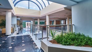 Un hall d’entrée spacieux avec un plafond haut et une verrière, doté d’un sol en marbre, d’un escalier central et d’un espace détente agrémenté de plantes.
