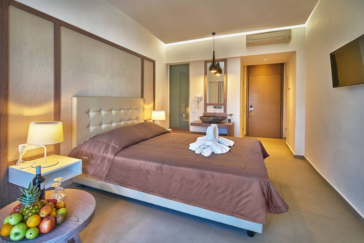 A modern hotel room with a large bed, bedside table with a lamp, a fruit bowl, and a TV mounted on the wall.