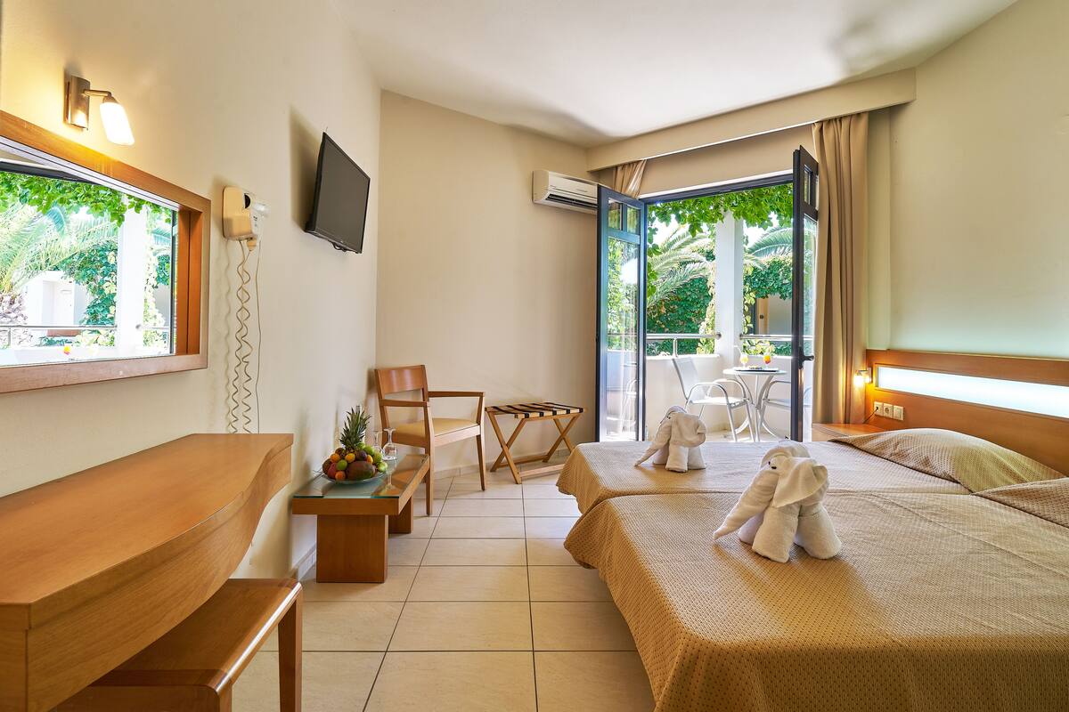 A hotel room with a wooden desk, two beds, a chair, a small table with fruit, and a view of greenery through the window.