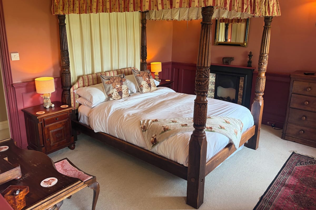 A four-poster bed with a canopy, a nightstand with a lamp, a dresser, and a mirror on the wall.