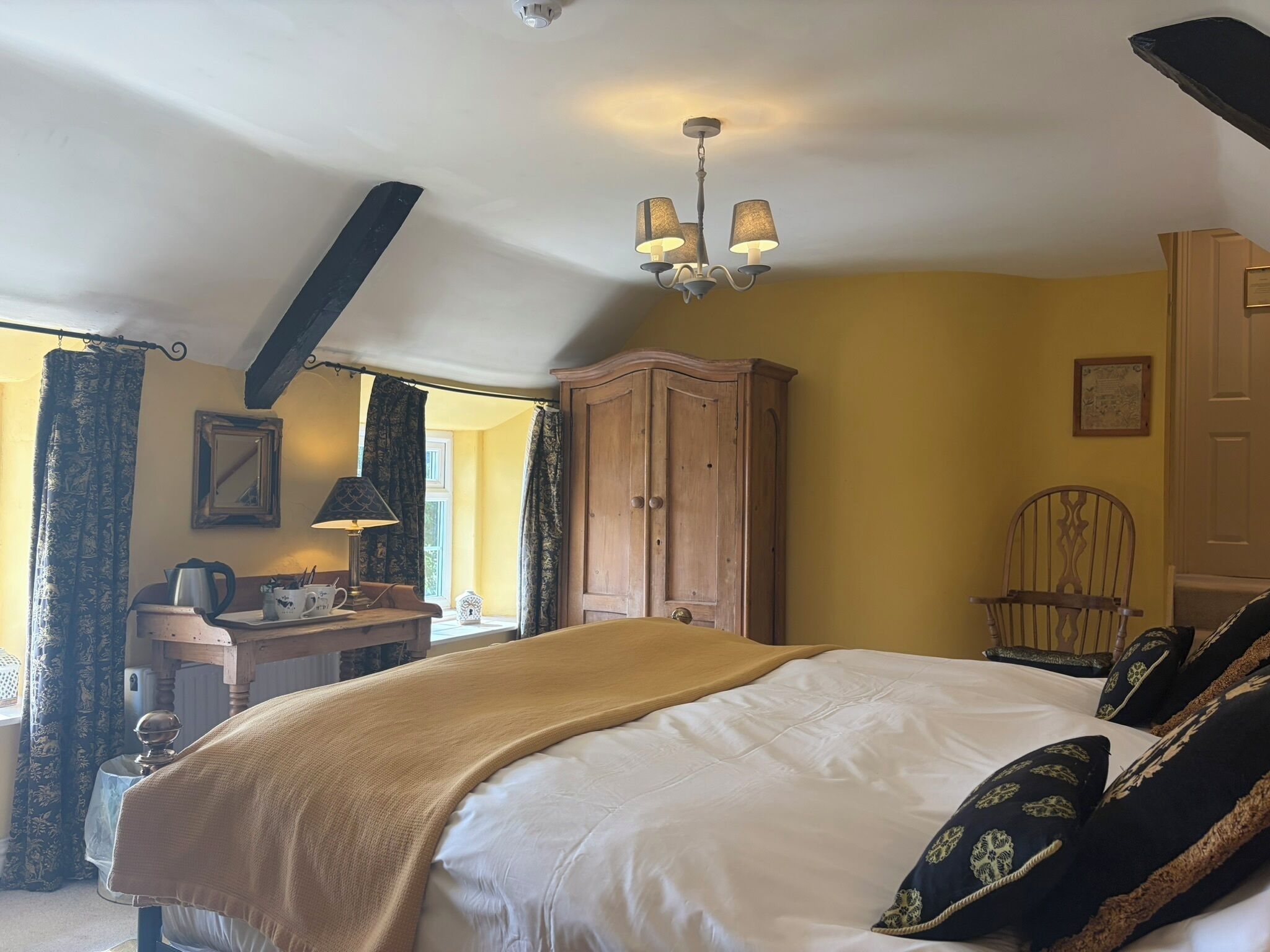 A bedroom with a bed, a wooden wardrobe, a desk with a lamp, a chair, and a window with curtains.