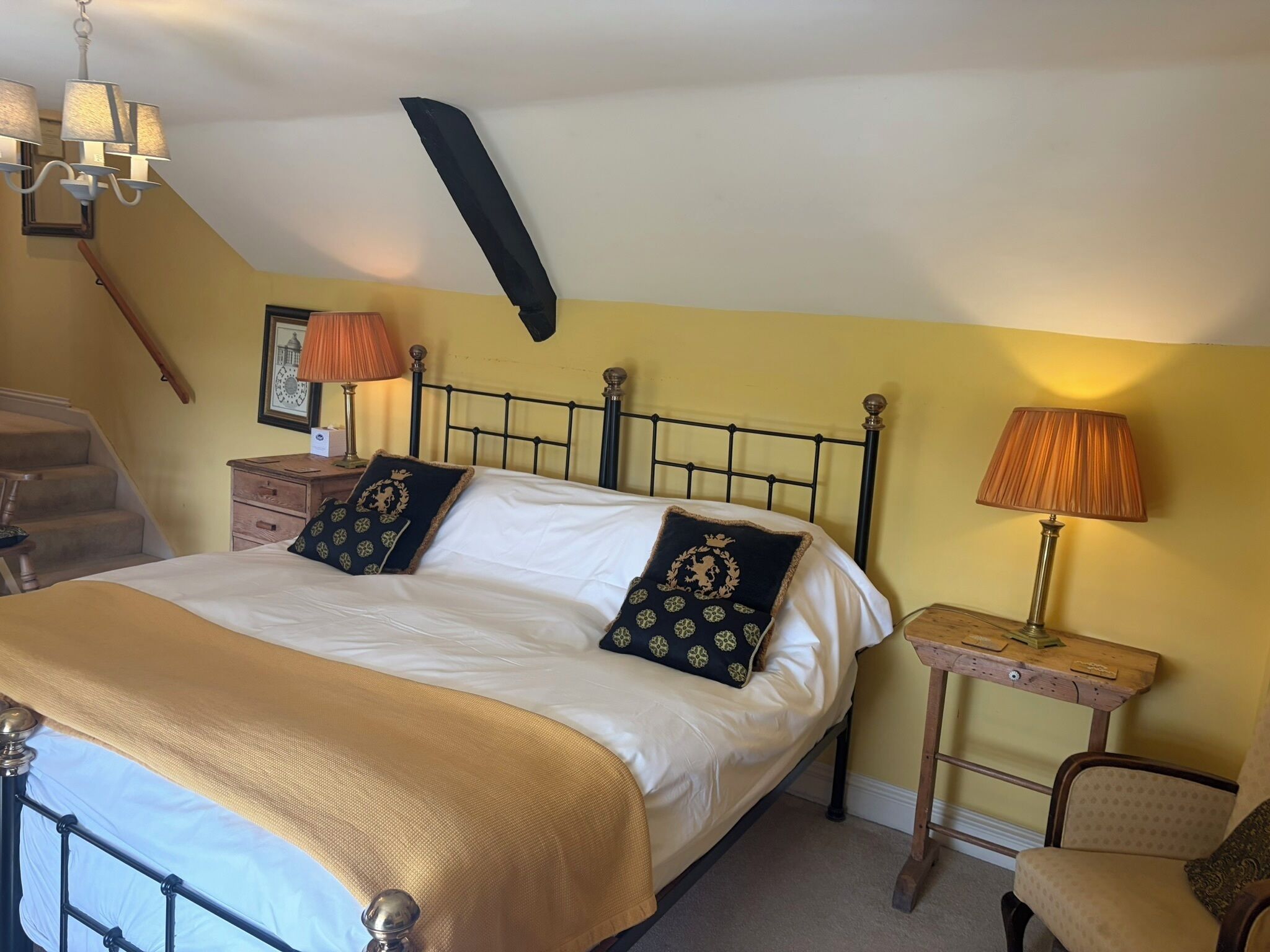 A bedroom with a bed, two bedside tables, a chair, a lamp, a picture frame, and a chandelier.