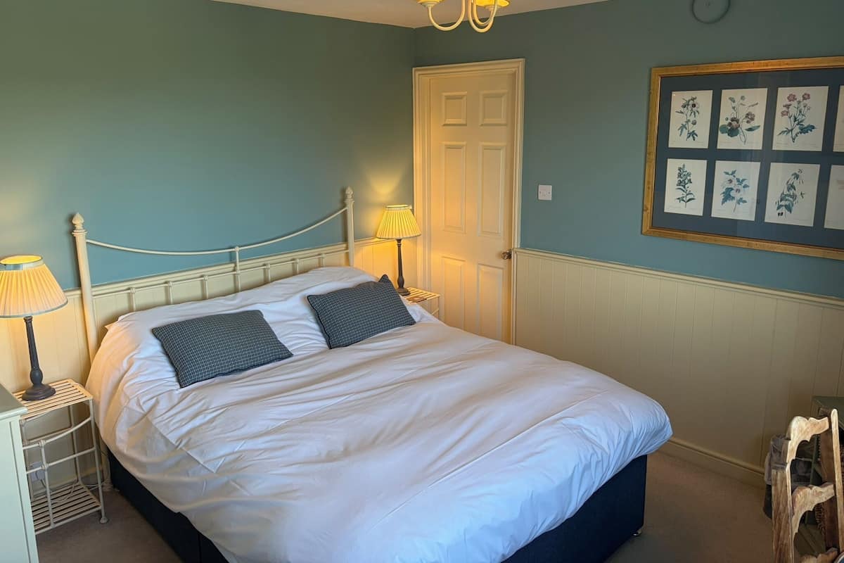 A neatly made bed with white linens and dark pillows, a bedside lamp, a framed botanical print on the wall, and a door with a white panel.