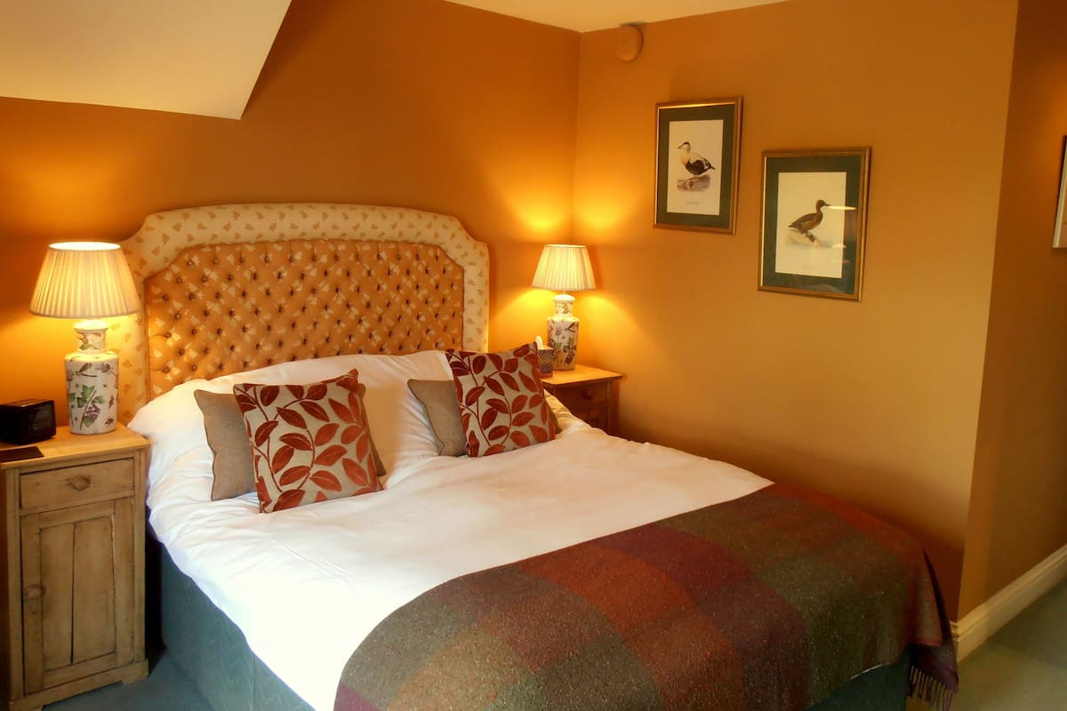 A bedroom with a tufted headboard, two bedside lamps, a bed with patterned pillows, and framed pictures on the wall.