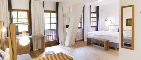 A modern hotel room with a wooden bed, a bedside table, a lamp, a wicker chair, and a large window with curtains.