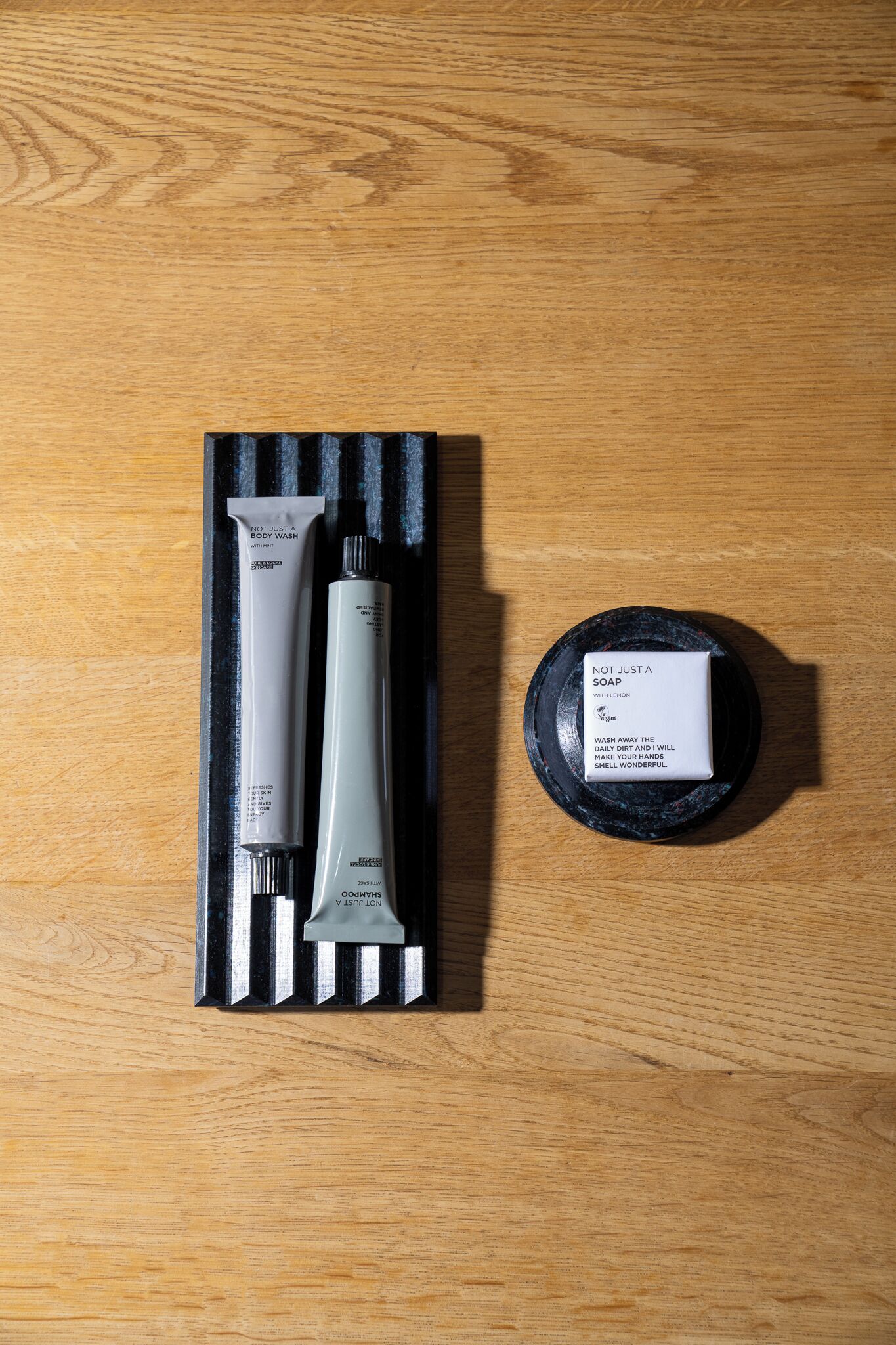 A wooden table with a black tray holding a set of personal care products including a shampoo, conditioner, and a soap bar.