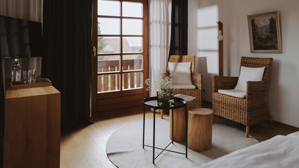 A hotel room with a large window, a wicker chair, a small round table, and a wooden nightstand.