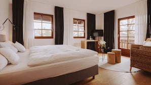A spacious bedroom with a large bed, two windows with curtains, a television, a small table with a vase, and a wicker chair.