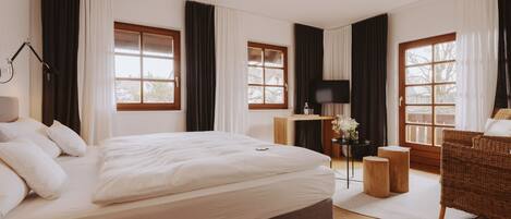 Un dormitorio amplio con una cama grande, dos ventanas con cortinas, un televisor, una mesita con un florero y una silla de mimbre.