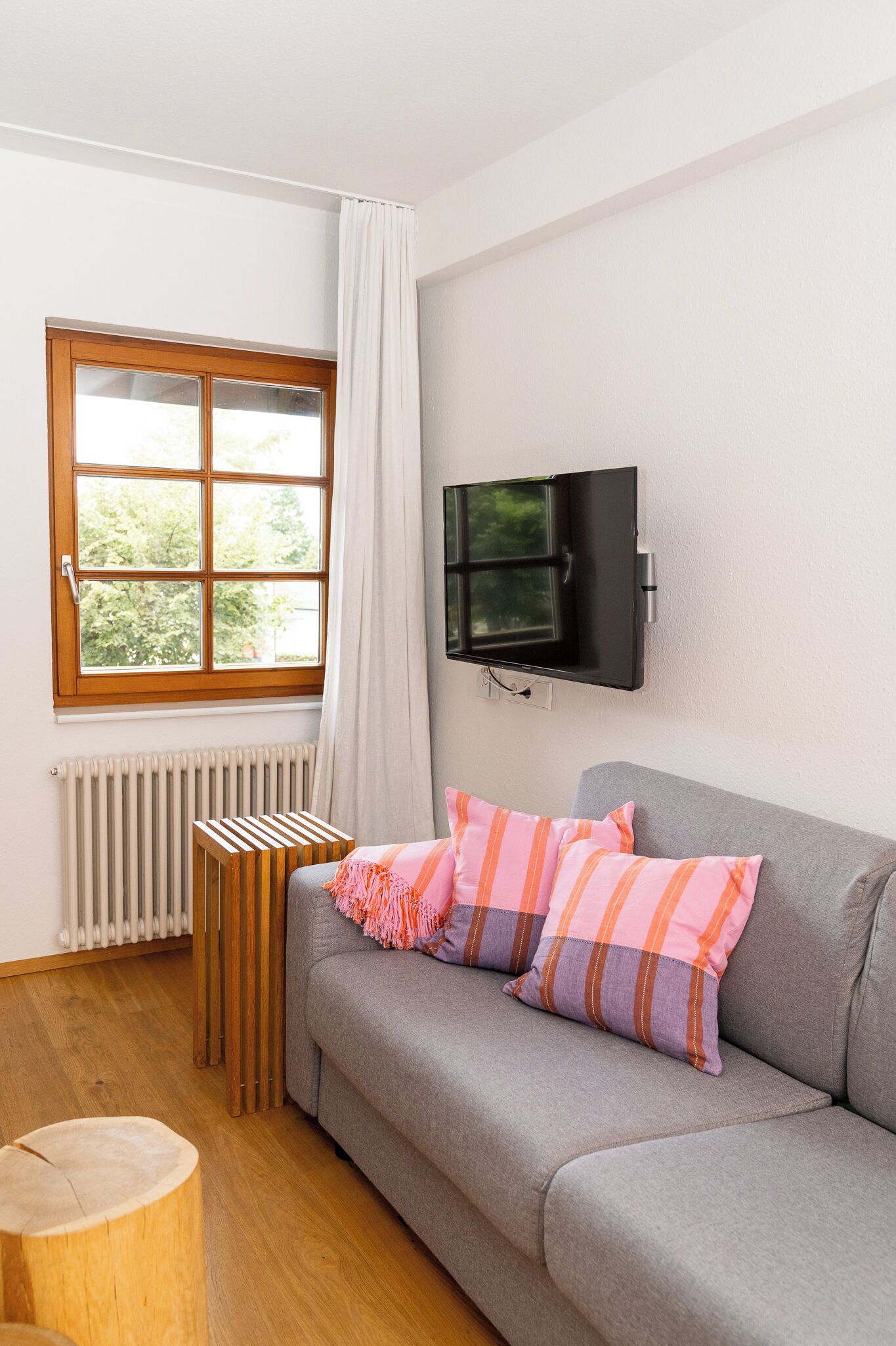 A modern living room with a grey sofa, a wooden radiator, a TV mounted on the wall, and a window with a white curtain.