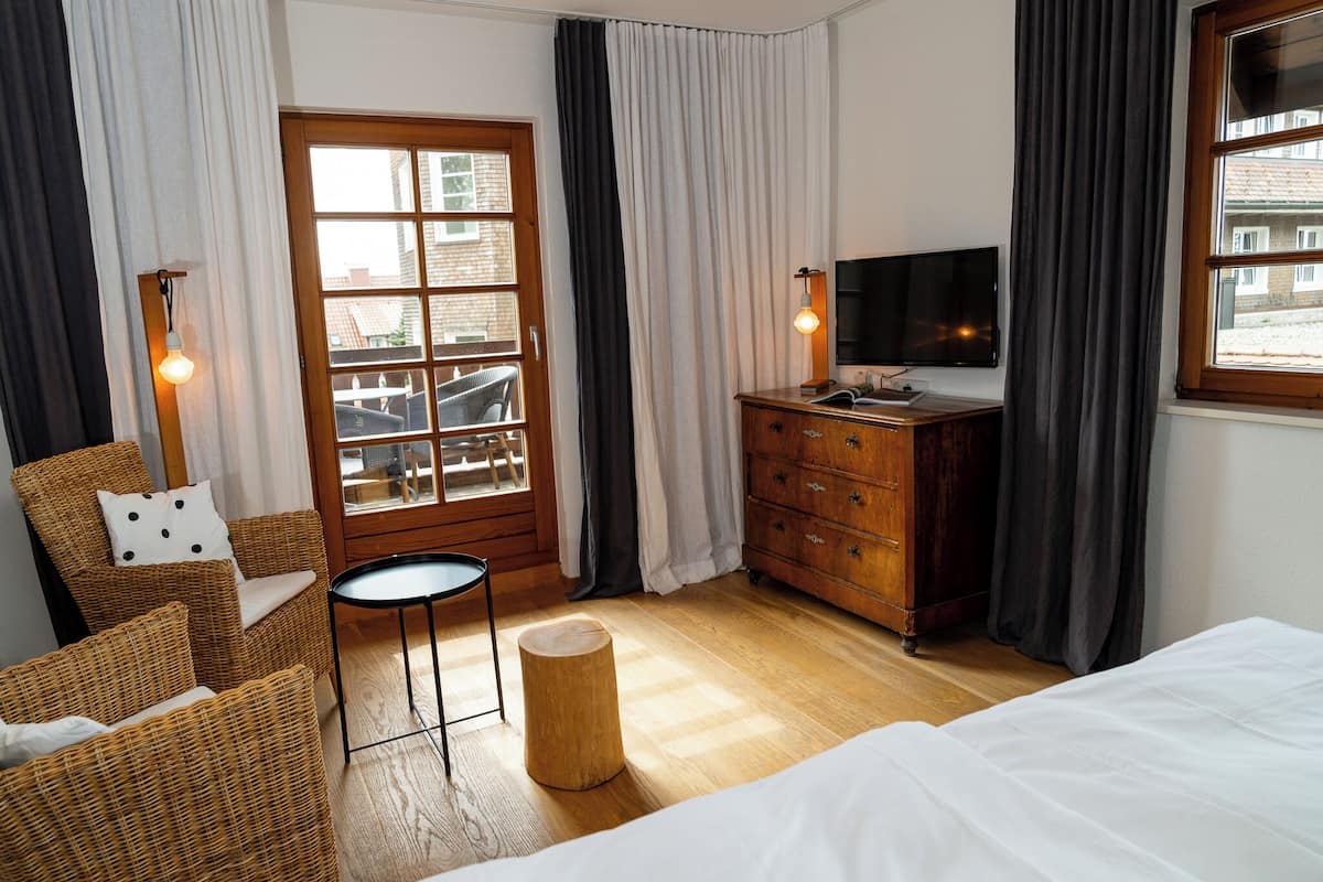 Habitación de hotel con una cama, cómoda de madera, televisor, sillas de mimbre, una mesita redonda y una ventana con cortinas.